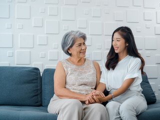 Portrait of happy Asian senior, mother or grandparent white hair holding hands of her beautiful daughter or grandchild smiling with love, care and comfort while sit on grey couch in living room.