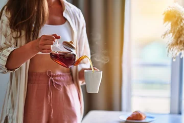 Raamstickers Woman pouring hot aromatic black tea into cup from glass teapot at home © Goffkein