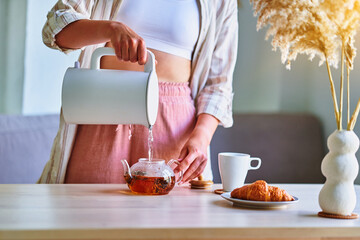Woman pouring boiling water into a cup from modern electric kettle for brewing tea at home