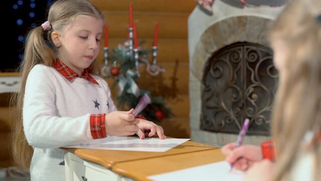 Little girls draw at the table near the fireplace and the Christmas tree. Two sisters write a letter to Santa Claus before the new year.