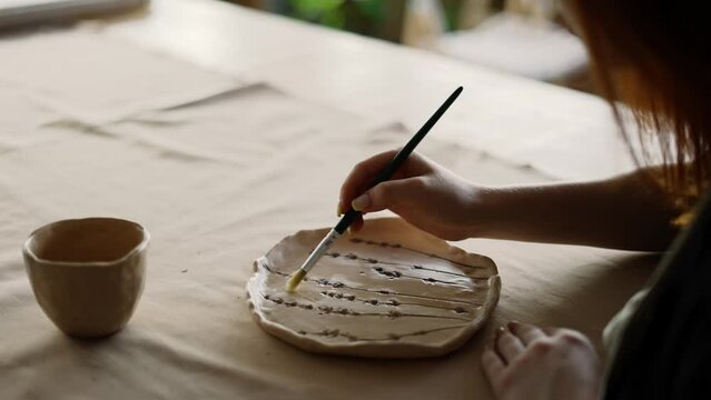 A red-haired girl potter varnishes a piece of clay with a special brush