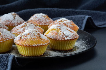 Freshly baked appetizing cupcakes sprinkled with powdered sugar on a black plate on a table with a dark surface