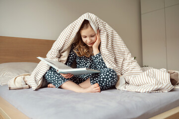 Cute teen child girl lying and reading book on bed under the blanket