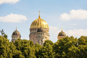 A church with a yellow wall and a pointed golden tip in the middle of the city center with multiple windows and trees next to it