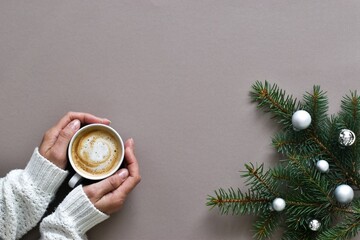 Winter drink. Female hands in knitted white sweater holding mug coffee and fir tree branch with balls on beige background. Flat lay creative composition. New Year and Merry Christmas concept 