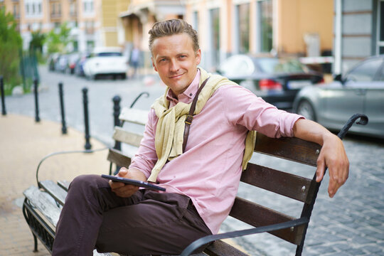 Freelancer young man with digital tablet in hands sits on the bench outdoors in front office building wearing casual pink shirt and shoulder bag. Young handsome man working on the go