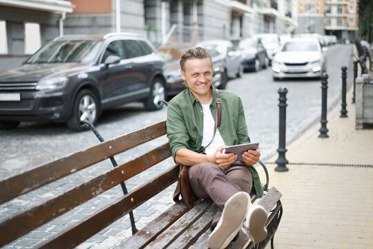 Happy freelancer man work using digital tablet sits on the bench outdoors in front office building wearing casual green shirt and shoulder bag. Young handsome man working on the go