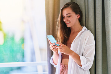 Satisfied carefree casual young beautiful happy smiling girl using phone at home interior