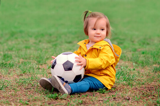 Little toddler girl in yellow jacket sitting on football field in summer or autumn day with soccer ball. Happy child playing football outdoors. Seasonal child activity