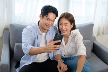 Obraz premium Young smiling asian couple using smartphone in living room at home