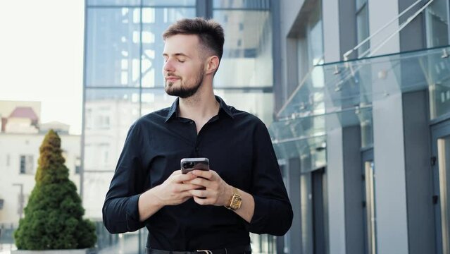 Young Businessman Walking near the Modern Glass Building. Looking at Smartphone Screen Messaging Online in Social Media Outdoor. Man is walking in Business Quartal, having good Mood looking Satisfied.
