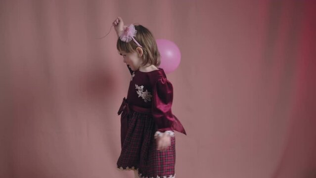 Cute little girl in an vintage burgundy dress is playing with a pink inflatable balloon. The concept of children theater productions and developing acting circles. Fuchsia color background
