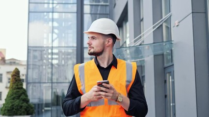 A handsome designer engineer in protective helmet and vest goes to work on the background of a modern building, uses the phone, examines the building project. Male professional reading email, message.
