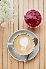 White ceramic cup of cappuccino with heart, raspberry jam dessert - a snack, light breakfast on the cafe terrace with blurred foreground of chamomile flowers. Flat lay food. Vertical photo