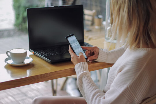 Side image of beautiful woman in striped shirt calling by smartphone while touching a white blank screen computer tablet with keyboard case and sitting at the wooden table with windows as background.