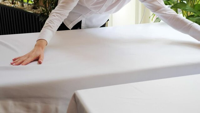 A female waiter spreads a white tablecloth on a table in restaurant against a background of green flowers. Preparation for celebrating event or a wedding. Catering, celebration, dining room, festive.