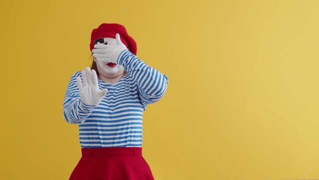 Clown girl in professional makeup emotionally protects her eyes with one hand and waving the other hand in stop gesture. Imaginary fear, fright, disgust or disapproval concept