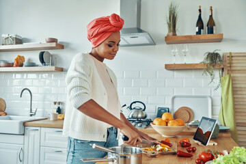 Confident young African woman in traditional headwear cutting veggies at the kitchen