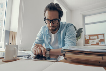 Handsome young man working on digital tablet while sitting at his working place in office