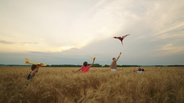 happy teens and baby boys are playing with toy planes and flying kite in gold rye field, slow motion