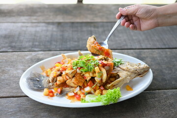 The right hand he holds a stainless steel spoon to scoop pieces of fried fish from a Thai dish called Deep Fried Sea Bass with Three Flavors Sauce. Fried fish with chilli sauce served in white plate.
