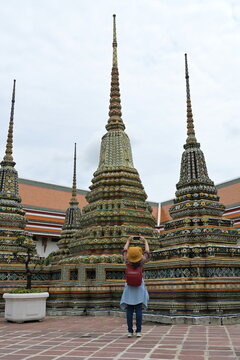 Woman traveler with wearing  yellow hat with red backpack sightseeing raise your mobile phone to take pictures of the  stuppas in Famous buddhist Temple :Wat Pho or Reclining Buddha Temple in Bangkok
