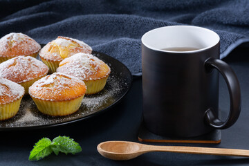Freshly baked appetizing cupcakes sprinkled with powdered sugar on a black plate and a cup of tea on a table with a dark surface
