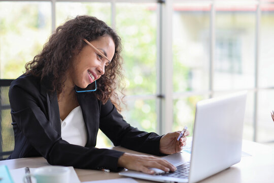 Attractive professional latin female employee worker sitting, using laptop computer with paperwork at home workplace. Businesswoman using smart mobile phone while holding credit card.