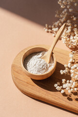 Wooden bowl with a spoon filled with white bath sea salt. Beauty treatment for spa and wellness on beige background. Skincare natural cosmetic concept for body care with dry plants and shadows