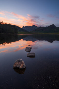 Beautiful sunset sky reflecting in perfectly still water with rocks in foreground and mountains in background. Blea Tarn, Lake District, UK.