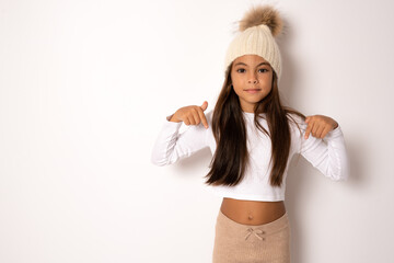 Pretty brunette little girl in winter hat pointing finger down isolated over whit background.
