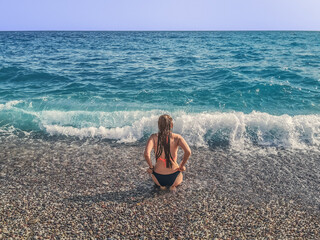 Young woman in a black bikini is squatting in front of a big wave on a pebble beach - view from the back. Adult girl in a swimsuit with wet hair on the shores of Mediterranean Sea in Antalya, Turkey