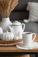 Still-life. Dried pampas grass in a vase, white ceramic pumpkins, a teapot, a cup and pumpkin-shaped candles on a coffee table in the home interior of the living room. Cozy autumn concept.