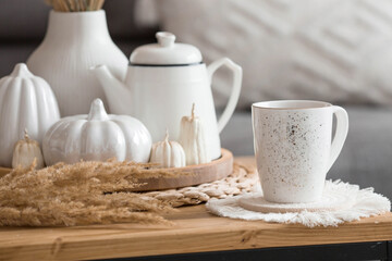 Still-life. Dried pampas grass in a vase, white ceramic pumpkins, a teapot, a cup and pumpkin-shaped candles on a coffee table in the home interior of the living room. Cozy autumn concept.