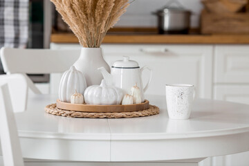 Still-life. Dried pampas grass in a vase, white ceramic pumpkins, a teapot and pumpkin-shaped candles on a white table in the interior of a Scandinavian-style home kitchen. Cozy autumn concept.