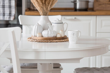 Still-life. Dried pampas grass in a vase, white ceramic pumpkins, a teapot and pumpkin-shaped candles on a white table in the interior of a Scandinavian-style home kitchen. Cozy autumn concept.