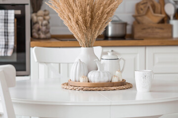 Still-life. Dried pampas grass in a vase, white ceramic pumpkins, a teapot and pumpkin-shaped candles on a white table in the interior of a Scandinavian-style home kitchen. Cozy autumn concept.