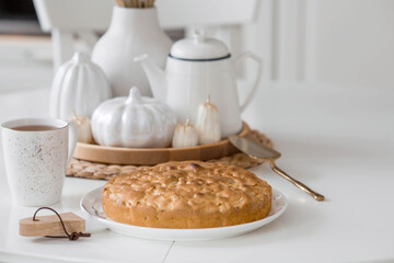 Still-life. Dried pampas grass in a vase, white ceramic pumpkins, candles and apple pie charlotte on a white table in the interior of a Scandinavian-style home kitchen. Cozy autumn concept.
