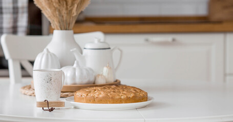 Still-life. Dried pampas grass in a vase, white ceramic pumpkins, candles and apple pie charlotte on a white table in the interior of a Scandinavian-style home kitchen. Cozy autumn concept.