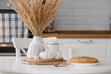 Obraz na płótnie Canvas Still-life. Dried pampas grass in a vase, white ceramic pumpkins, candles and apple pie charlotte on a white table in the interior of a Scandinavian-style home kitchen. Cozy autumn concept.