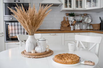 Still-life. Dried pampas grass in a vase, white ceramic pumpkins, candles and apple pie charlotte on a white table in the interior of a Scandinavian-style home kitchen. Cozy autumn concept.