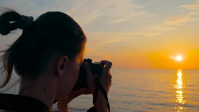 Woman is standing on the sea shore, holding photo camera and taking photo or shooting video of the Black Sea, warm sunset sky - close up, slow motion, sun lens flares. Photography and summer concept