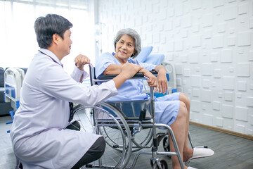 Asian doctor supporting and cheering up senior patient in wheelchair talking, smiling in comfort at hospital. Healthcare and medicine concept.