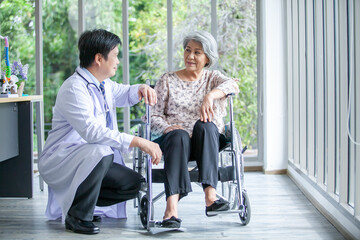 Fototapeta na wymiar Asian doctor supporting and cheering up senior patient in wheelchair talking, smiling in comfort at home. Healthcare and medicine concept.