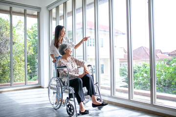 Beautiful asian girl help and take care of senior elderly woman sitting on wheelchair at living room in house, senior therapy patient at home concept. - 531462378