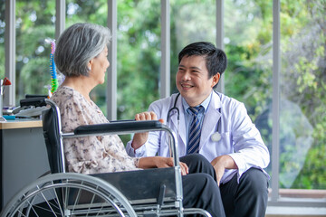 Asian doctor supporting and cheering up senior patient in wheelchair talking, smiling in comfort at home. Healthcare and medicine concept.