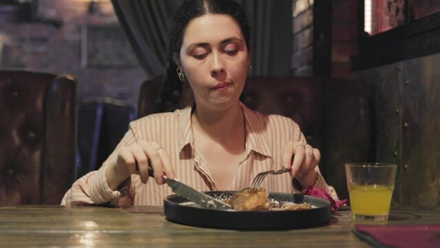 A young Caucasian pretty woman holds cutlery in her hands and unsuccessfully tries to cut off a piece of fried fish. Dinner at the restaurant.