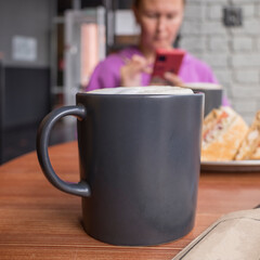 a black coffee mug on the table in a cafe against the background of a woman with a smartphone