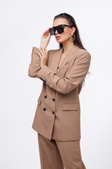Fashion photo of a beautiful elegant young woman in a pretty oversize brown and beige suit, jacket, pants, stylish sunglasses posing over white background. The hair is gathered back, dark brunette.