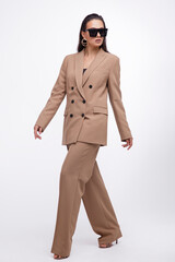 Fashion photo of a beautiful elegant young woman in a pretty oversize brown and beige suit, jacket, pants, stylish sunglasses posing over white background. The hair is gathered back, dark brunette.